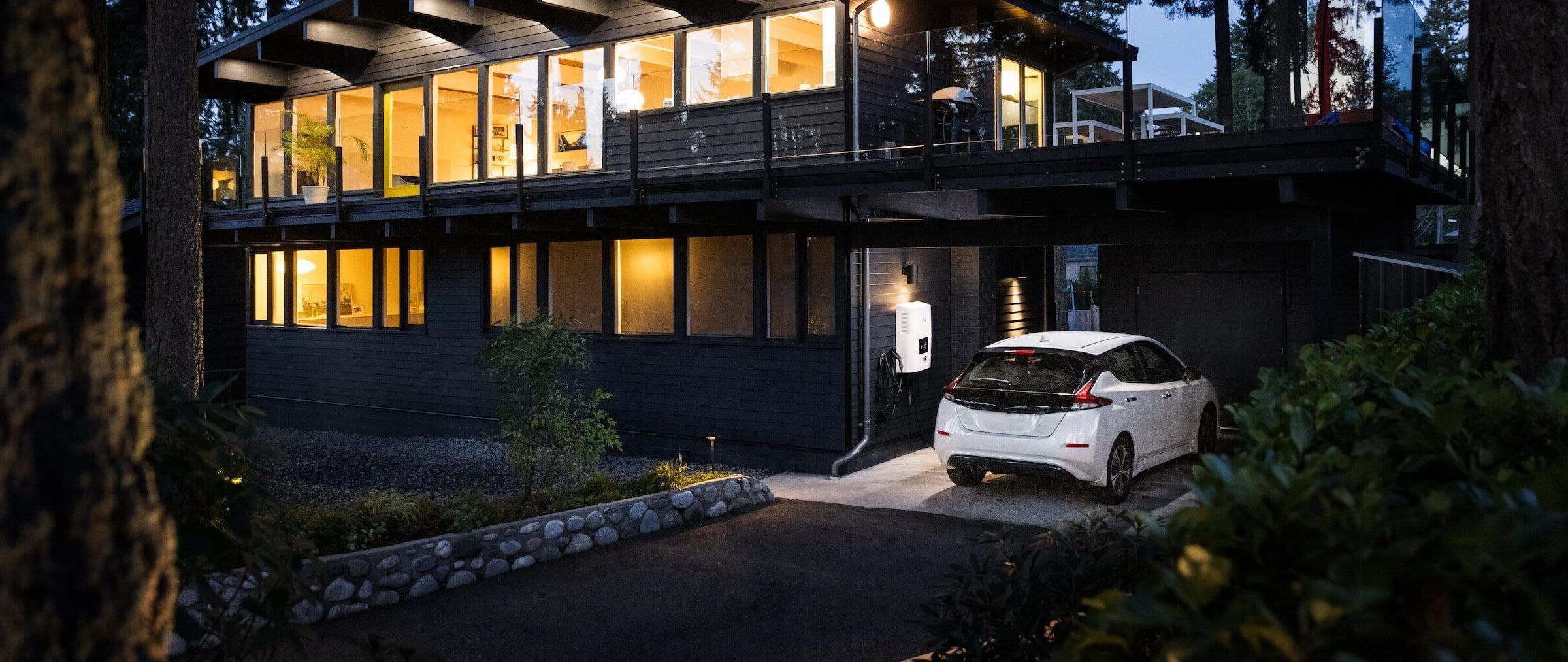 Ein Elektroauto steht in einer Auffahrt an einer Wallbox neben einem durch Öko Strom beleuchteten Haus